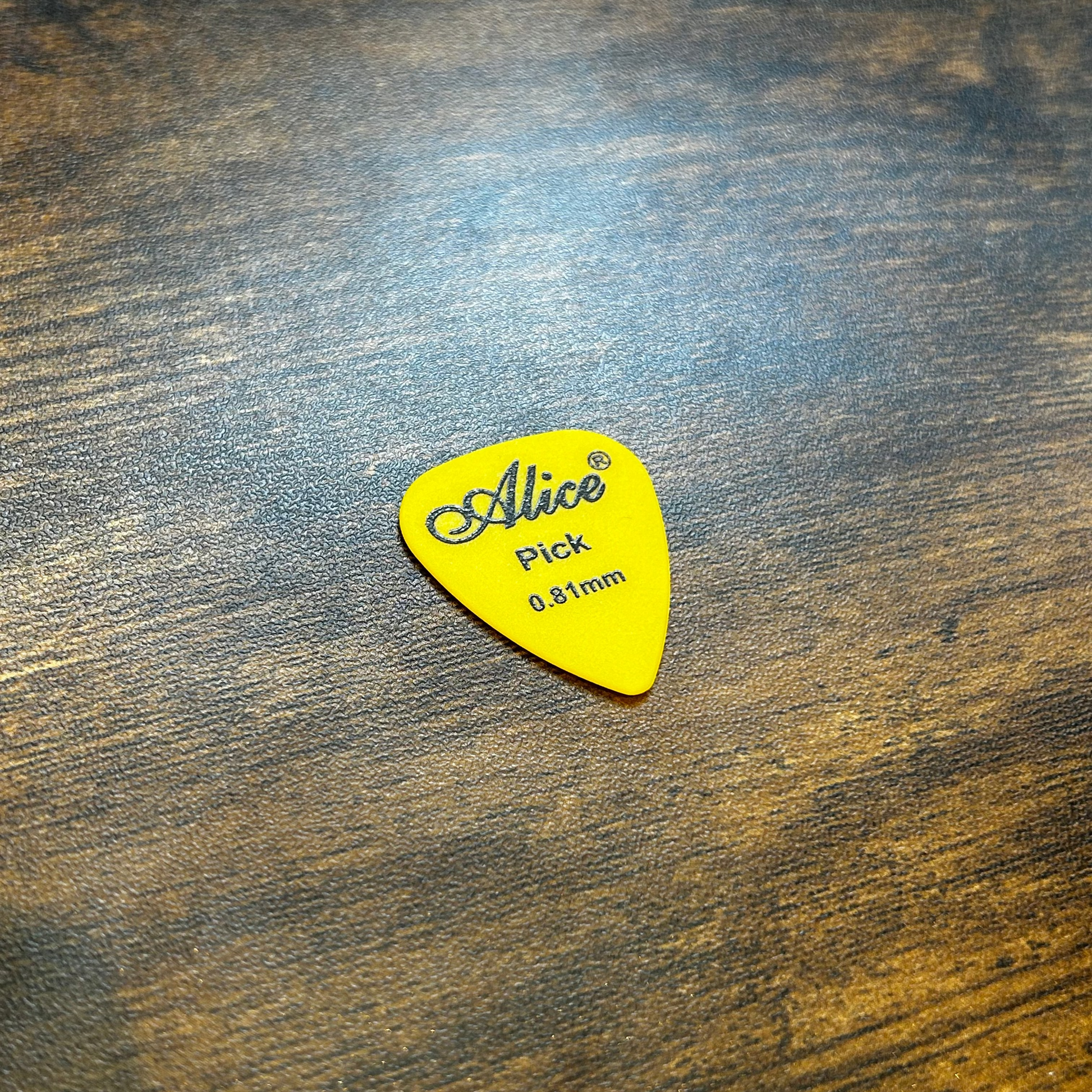 Yellow 0.81mm guitar pick with 'Alice' branding on a wooden surface