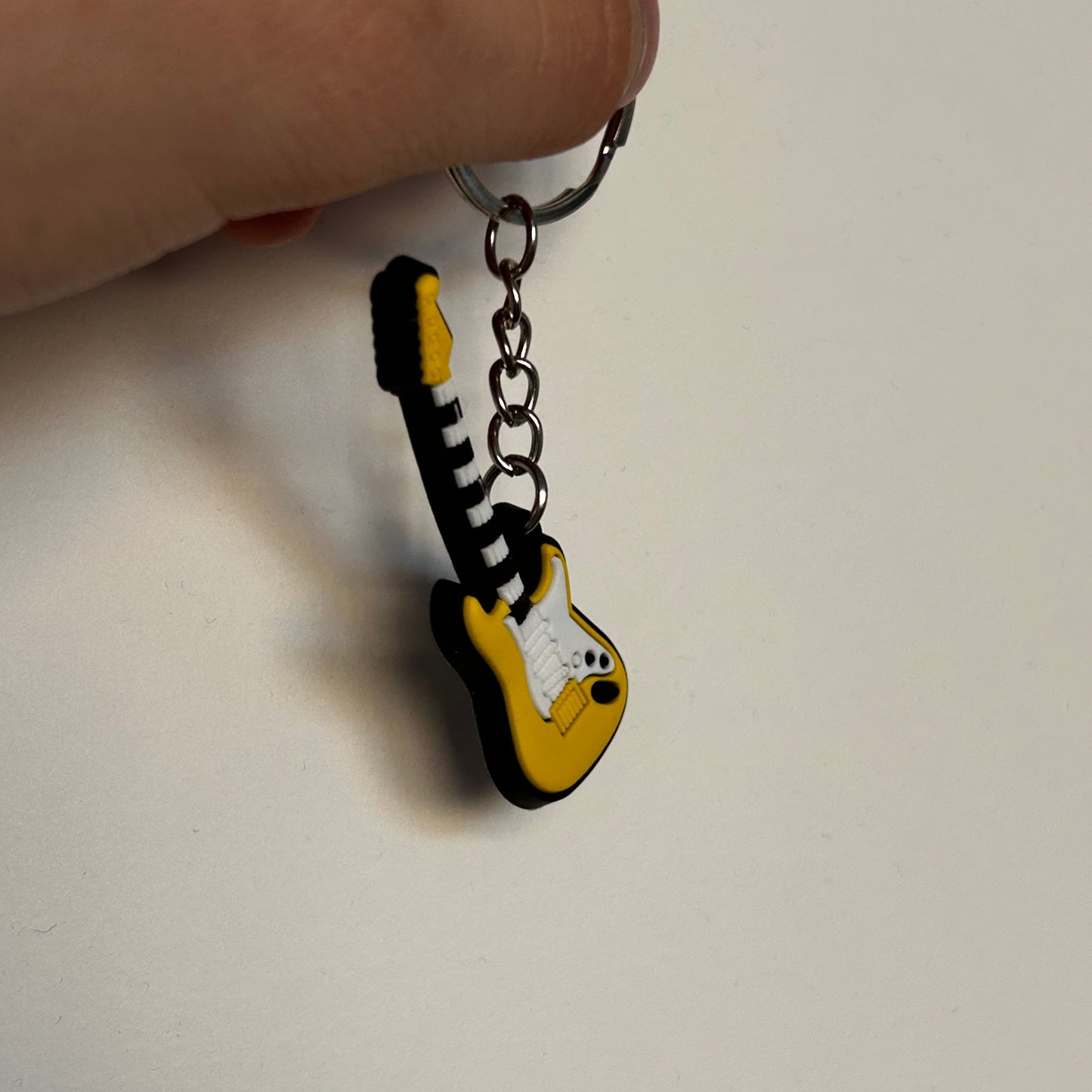 A small guitar-shaped keychain held by a hand against a plain off-white background. The keychain features a yellow and black electric guitar design with a silver metal keyring.