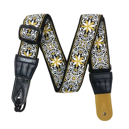 A guitar strap with a black, white, and gold patterned fabric body, black leather hardware, and a tan leather end piece, displayed on a white background.