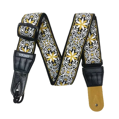 A guitar strap with a black, white, and gold patterned fabric body, black leather hardware, and a tan leather end piece, displayed on a white background.