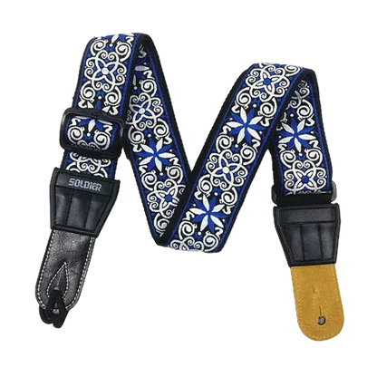 A guitar strap with a blue and white ornate scroll pattern, black leather ends, and a gold-toned metal hardware piece, displayed against a solid black background.